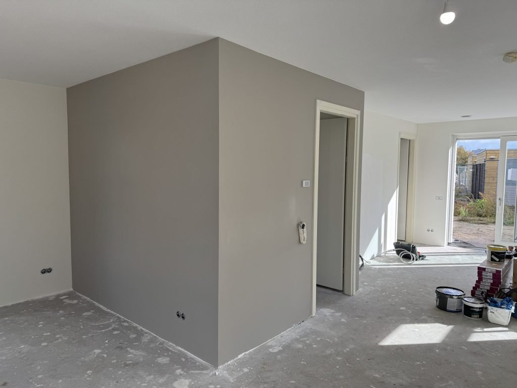 Vrijstaande wand in een grijze tint naast een deuropening, met verfemmers op de vloer