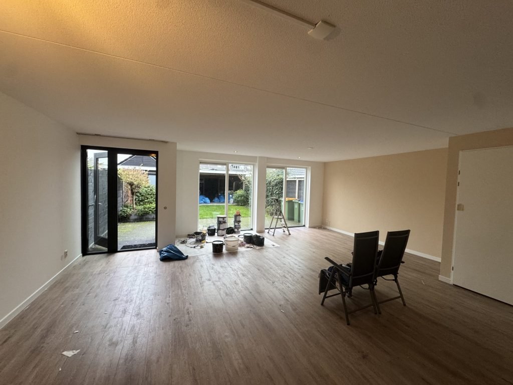 Ruime woonkamer met lichte wanden, een houten vloer en schildersmateriaal bij de tuindeuren